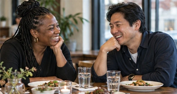 A Black woman and an East Asian man laughing together over dinner